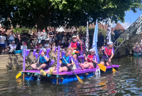 A group of people in life jackets are sitting on a purple and blue raft, floating on a river. There is a crowd of people on the riverbank.