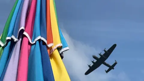 if all_else A multi-coloured parasol can be seen on the left-hand side of the photo with a bright blue sky from above showing on the other side. In the sky is an aircraft presumably part of the Bournemouth Air Festival last week.