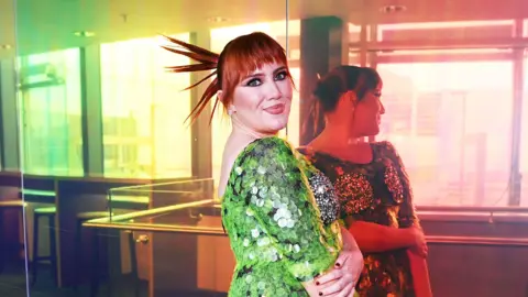 CMAT, wearing a scaled green dress, posing against a reflective multicolour window