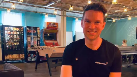 Mousumi Bakshi/BBC Image shows man in a black tee shirt looking into the camera with arcade machines behind him.