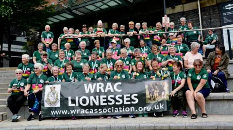 Handout Dozens of runners wearing green tops are sitting on steps. The front row is holding a banner which reads: WRAC Lionesses