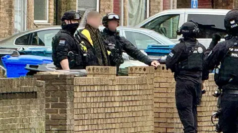 A man is led out of a house by two armed police officers in black tactical gear. The man is wearing a black and yellow jacket and has short brown hair.
