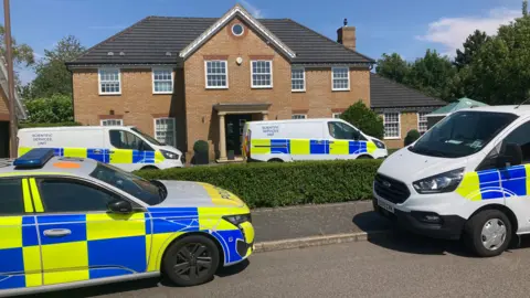 DJ McLaren/BBC A police car and a few forensic vans are parked outside a large house, some on the street, some on the driveway