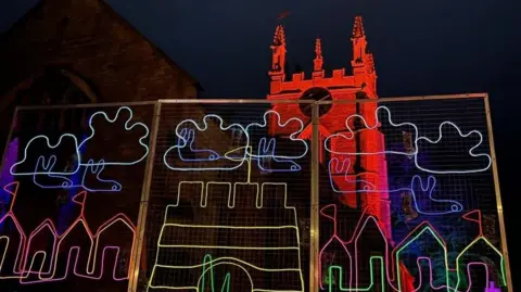 East Lindsey District Council Illuminated artwork in front of a church featuring clouds and a yellow-coloured sandcastle 