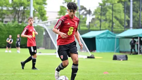 FAW A boy with dark curly hair, wearing a red Wales football shirt, black shorts, black football socks and white trainers. He is kicking a football on a green grass football field, with other players and a goal behind.