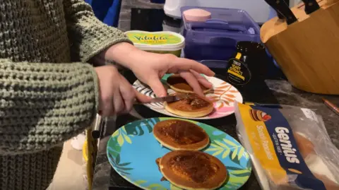 Alex Osborne/ BBC Image shows Debbie making the childrens' breakfast; two colourful plates topped with small, round pancakes, spread with marmite.