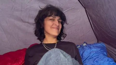 Henrietta Henrietta, a woman with short brown hair and a fringe, smiles. She is sitting down in a tent and is wearing a long-sleeved black top and two necklaces around her neck.