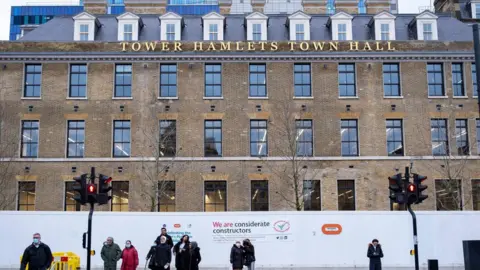 Getty Images An image of Tower Hamlets Town Hall, a brown brick building with white hoardings on the ground floor. The view shows people waiting at a traffic light ready to cross, and in the foreground there is a road,