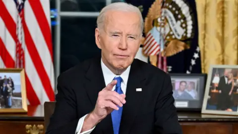 Joe Biden sits astatine  a table  with photos and a US emblem  down  him and points towards the camera.