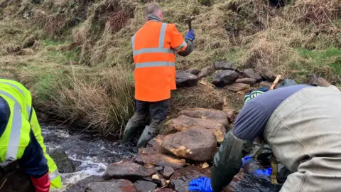 Nicola Rees/BBC Three mean wearing waterproof gear stood in a stream building a wall.