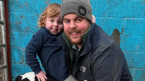 Andrew Little and daughter Molly in navy boiler suits, smiling holding a black and white calf, pictured against a bright blue wall in a farm shed