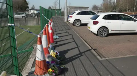 Six traffic bolllards in front of a gap in some green mesh fencing with bunches of flowers laid in front of them. A car park is to the right, with two white vehicles parked up.