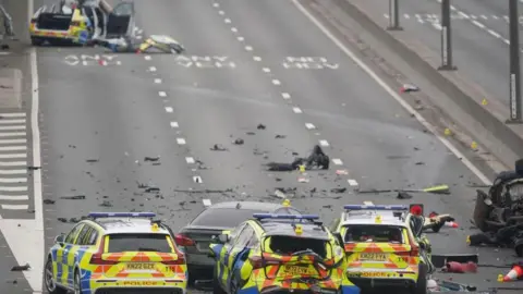 PA Media Close-up photo of three police cars with debris on the road. One of the cars has its rear massively damaged and crushed. They surround a grey car. A fourth police car in the background has its roof taken off.
