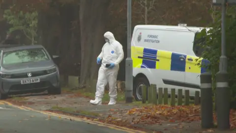 A forensic investigation officer wears a white suit that covers their head and feet, with blue gloves. They are standing next to a Staffordshire Police forensic invest van with blue and yellow livery