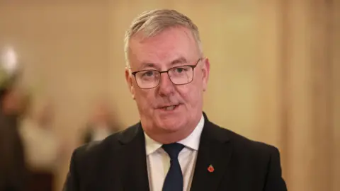 PA Mr Nesbitt wears glasses and a black suit jacket, white shirt and navy tie as he talks to the camera.