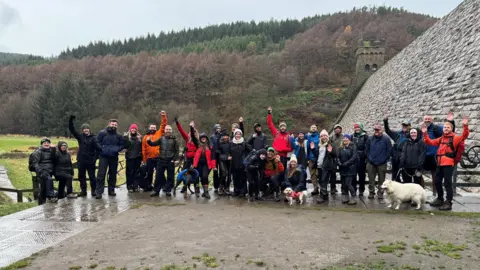 A group of 28 walkers and some dogs cheering in rainy conditions at Derwent Reservoir in Derbyshire 