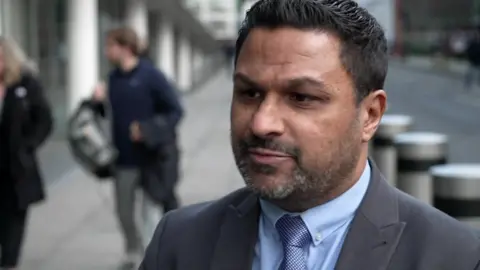 Andy George is seen wearing a suit being interviewed on the street. Others can be seen out of focus in the background.