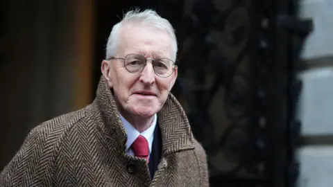 PA Media Hilary Benn is wear glasses and his wispy white hair is being blown upward by the wind. He has on a heavy tweed jacket with high-collars and his white shirt-collar and red tie can protrude from the unbuttoned neck of his coat. 