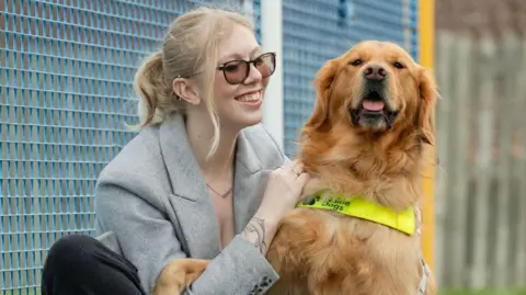 Hannah Burgess is wearing a grey jacket and crouched down holding the harness of Morris, a golden retriever. Hannah is looking at Morris. Morris is looking at the camera. Morris has a yellow high visibility harness on. Hannah is smiling. They are standing in front of a blue metal fence, which appears to be part of a children's play area or sports facility. A yellow pole and another metal fence can be seen in the background. 
