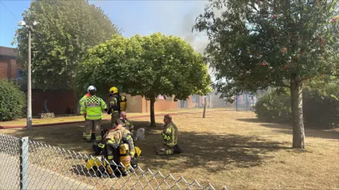 Fire fighters sit on parched ground inside Thomas Bullock Primary Academy. In the distance is smoke from a fire in a school building. Trees are on the field. 