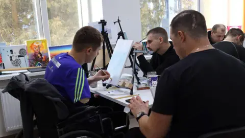 Veterans' House A man with his right arm missing sits in a wheelchair painting. He is wearing a blue and green football shirt. 