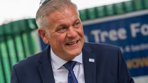 Marlon West, who has grey hair and is wearing a navy blue suit over a white shirt and blue tie, smiles into the camera. 