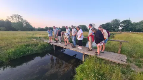 A group of about a dozen people standing on a small wooden bridge over a canal looking at what's in the water. The bridge goes between two grassy fields. It appears to be around dusk, with a gold light shining on the ground.