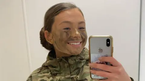 PA Media A mirror selfie of Jaysley Beck wearing a camoflauge jacket with green and brown face paint on her face. Her brown hair is tied up in a slick back low bun and she is smiling at the camera.