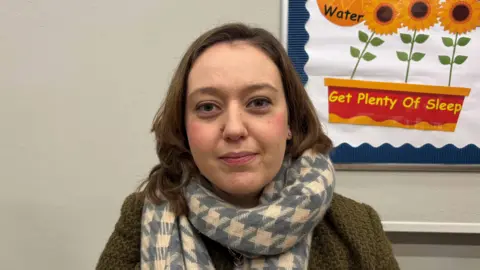 A woman with long brain hair looks at the camera. She is wearing a green cardigan and a blue and white scarf.