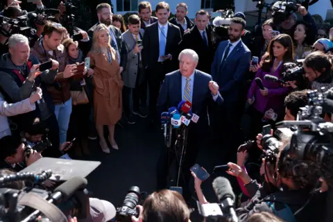 Senator Graham in a suit and tie is surrounded by reporters outside the White House. He speaks into a microphone with his hand raised emphatically