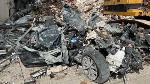 Burnt and gnarled metal, with tires visible, is in a pile on the ground, with a CAT machine in the background.