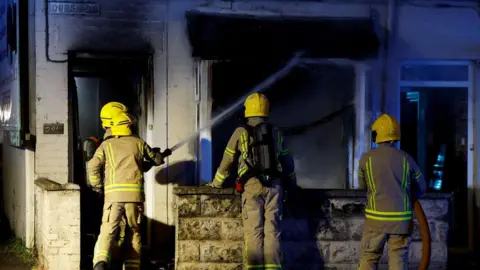 Reuters Firefighters working to put out a fire in a house during the riots. They are wearing their uniform with yellow helmets. There are four of them in the picture.