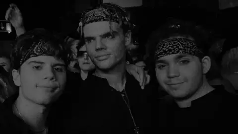 Lincolnshire Police A black and white photo of three young men wearing patterned bandanas and smiling together into the camera. They have short hair and the man in the middle is taller and has braids in his hair. There are several people behind them in an enclosed space.
