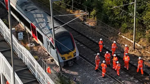 PA Media Part of a Greater Anglia train service derailed at Wickford in Essex. A number of people wearing fluorescent orange outfits with white helmets are stood on the tracks near the train.