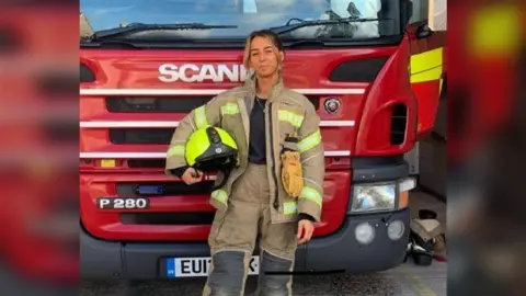 A woman in firefighter gear standing in front of a red fire engine.