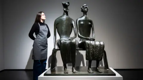A bronze sculpture of a couple sitting on a bench. A woman in a black top and grey apron stands next to the sculpture with her hands behind her back, looking at it.