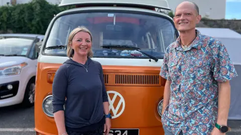 Alyson Grainge and Sebastian Wale pictured smiling stood in front of an orang VW campervan. Alyson is wearing a navy hooded top and has shoulder-length blonde hair. Sebastian is wearing a patterned button down shirt.