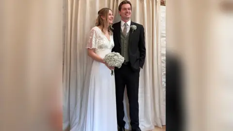 Rebecca and George Lance Rebecca wearing a floor-length white wedding dress and holding a bouquet of flowers, stood next to George who is wearing a three-piece suit with green tie 