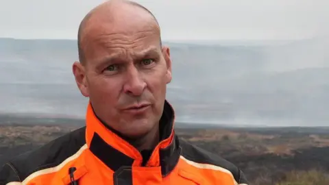 Oli Constable/BBC Andy Creasey wearing an orange and black coat, looking serious into the camera, with a shaved head. He is stood where the fire has been burning and there is smoke in the background over the heather and fields.