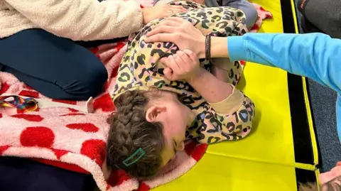 Liz Chaplin Ava is wearing a colorful leopard print jumper. She is lying on her side during a physiotherapy session and is being assisted by two people.