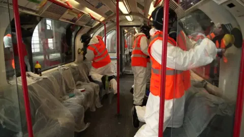 BBC Three men dressed in white protective overalls and hi viz vests clean the graffiti inside a train with buffers