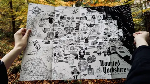 A person holds a map titled Haunted Yorkshire in a forest. The map features playful illustrations of aliens, giants, fairies and ghosts attached to location names. 