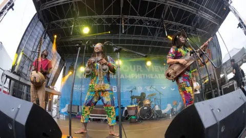 Bristol Harbour Festival An image of a group of three musicians wearing brightly coloured clothing and playing traditional instruments on a festival stage. 