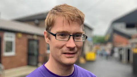Elizabeth Baines/BBC Kevin Darby - a man with short ginger hair and glasses - looks into the camera. He is wearing a purple T-shirt.