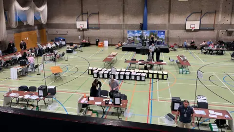 Image of an election count in Huntingdonshire. It is in a sports hall with ballot boxes lying around and tables where people are set to council and adjudicate.