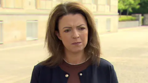A woman is standing outside in front of a large building. She has shoulder length brown hair. She's wearing a brown jumper and a navy jacket.