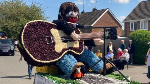 One of the floats is a depiction of a man with a guitar, singing at a microphone. He has brown hair, glasses, red lips and is wearing blue jeans.