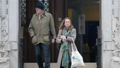 A woman and a man coming down steps outside Newport Crown Court. The man, on the left, wears dark blue trousers and a khaki coat, and a dark blue woolly hat. The woman, on the right, wears a grey coat and green patterned scarf; she carries a tote bag.