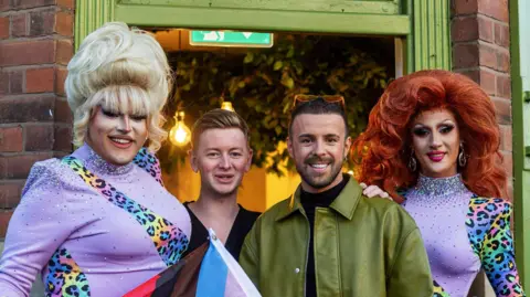 The Secret Garden Four people stood in the salon door way. A drag queen with blonde hair and a drag queen with red hair stand at each side of two men in the middle. 