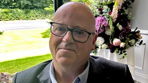 BBC Mark McCollum wearing a blue shirt, blazer and glasses. He is looking at the camera.